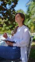 Young woman engaging with digital tablet while sitting on park bench, showcasing dynamic expressions and interactions amid natural, sunlit environment during leisure moments video