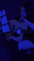 Woman musician with guitar sits on bar stool near music stand, leaf through notes and then begins to play guitar under flicker of multi colored neon lights in nightclub. Low light effect handheld shot video
