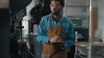 African American man working in roasted coffee fabric engineer technologist inspecting checking part of industrial roasting machine writing notes go away male roaster guy industry check equipment video