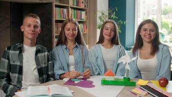 Four happy smiling looking at camera teenagers group classmates with paper craft house windmill project learning art therapy education friendship Caucasian pupils students together in school library video