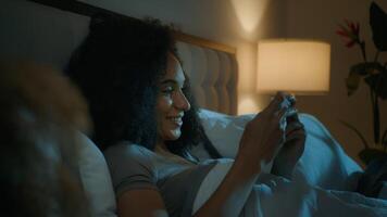 African American woman sleepless girl happy insomniac teen playing online game illuminated with mobile phone screen on bed in night gaming leisure cybersport fun playing gadget addiction before sleep video
