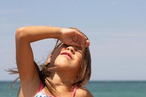 Beautiful caucasian child of 7-8 years looking at sun and covering face with hand on sea background photo