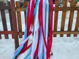 Multicolored white, red, and blue ribbons flutter in the snow on a wooden fence photo