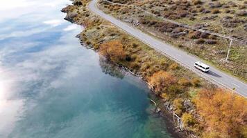 Drone captures a serene lakeside road in Cromwell, New Zealand, with autumn foliage and calm waters reflecting the sky video