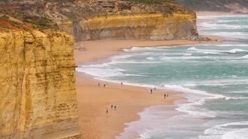 Tourists stroll along the sandy beach beneath towering cliffs at the Twelve Apostles, with waves crashing under overcast skies video