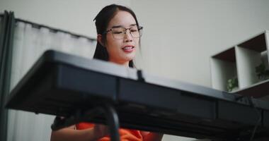 Handheld low angle view shot, Beautiful Asian woman playing the keyboard with happy smile in cozy living room. Enjoy a relaxing weekend activity video