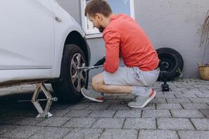 Close up of man changing a car tire in front of his house. The vehicle is lifted with a jack, tools lie nearby. The scene captures a hands-on repair moment in daylight photo