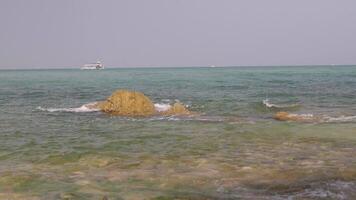 A view of the sea below eye level with waves washing over rocks and a tourist boat passing by. video