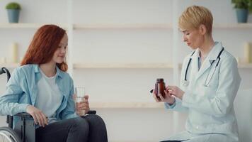 A healthcare professional wearing a lab coat explains medication to a girl sitting in a wheelchair while holding a glass of water. The setting is bright and welcoming. video