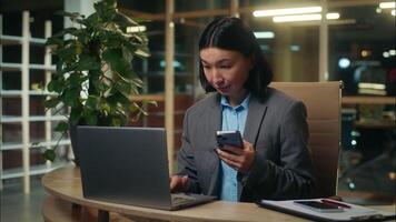 Asian Korean Chinese Japanese woman multitasking working using mobile phone smartphone laptop female girl businesswoman business office indoors manager employee online worker typing computer chatting video