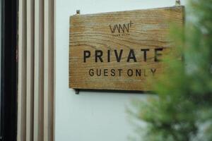 The wooden signboard with the words Private Guest Only hanging on the white wall with blurred tree in foreground photo