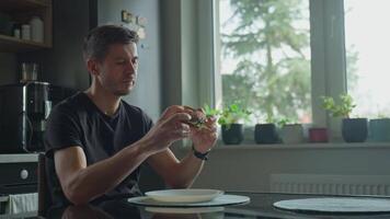 Man sitting at kitchen table eating sesame bagel with egg, cheese and ham. Person has breakfast with homemade sandwich video