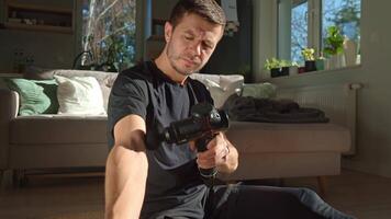 Man using percussion massage device. Man applies massage gun to arm muscle while sitting in living room, recovery therapy after exercise. Concept of home fitness and relaxation video