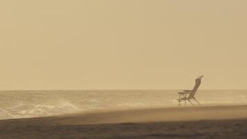Wind sweeping sand across desolate beach, lone chair casting solitary silhouette amid misty, atmospheric coastal landscape, generating sense of profound solitude and tranquil isolation video
