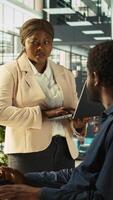 Vertical Worker in office receiving feedback from management executive looking over work done on laptop. African american manager in workspace giving tasks to employee, providing necessary data, camera B video