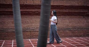 Handheld side shot, Solo female tourist enjoys listening to music from wireless headphones and playing smartphone while walking around the ancient site. Traveling and vacation concept video