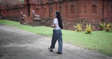 Handheld back view shot, Solo Asian tourist enjoys taking photos with camera while taking a leisurely stroll amidst serene surroundings and the cultural richness of the place. video