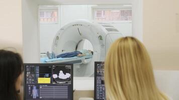 Two qualified radiologists examines female patient who is lying under CT or MRI scanner. Computer screen with result diagnosis image. View through glass window in a modern clinic. 2 video