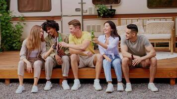 A group of diverse friends relaxes on a deck, enjoying cold drinks and laughter. They are sitting together, taking a break from camping activities on a sunny weekend. video