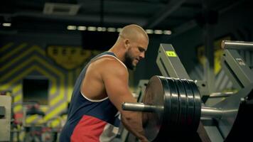 Strong bald man beard focuses on leg exercises while lifting weights in well equipped gym. hangs weights on leg press machine video