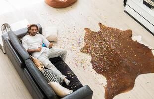 Young man relaxing on sofa with confetti chaos on floor in living room. Top view of home interior after celebration party. photo