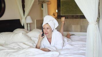 Relaxing woman in bathrobe enjoys phone conversation on cozy bed in a serene bedroom setting video