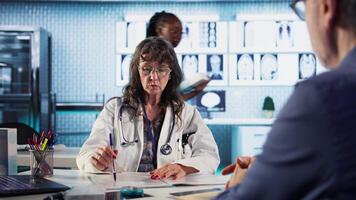 Senior doctor and patient discussing diagnostic results in a modern medical office, showcasing advanced technology using records files displaying x rays and MRI scans. Care management. Camera A. video