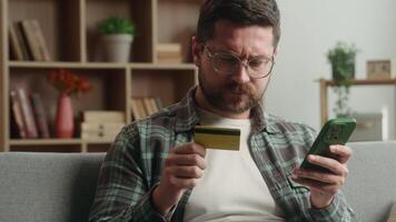 Adult man male buyer client at home living room using blocked credit card and smartphone for shopping on web stores purchase mobile phone fail error typing on cellphone trying to pay annoyed payment video