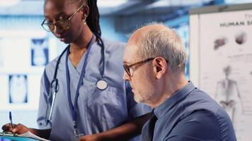 Expert and african american assistant provide medical services to patient at checkup, giving diagnostics report and medical records. Medical staff ensure healthcare exam on insurance. Camera B. video