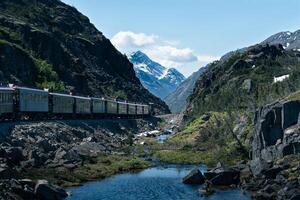 Train cars traveling through an Alaskan mountain pass along a stream of water with snowy peaks in the background and a blue sky with white clouds in the distance. photo