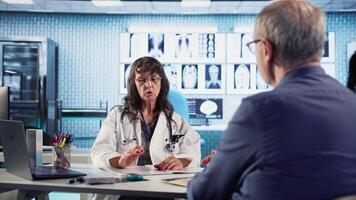 Senior woman evaluating diagnostic results with her medic in a cabinet, using advanced tech tools to give the correct results to his patient. Care management within medical industry. Camera A. video