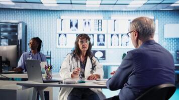 mayor médico y paciente que se discute diagnóstico resultados en moderno médico oficina, exhibiendo avanzado tecnología utilizando computadora mostrando X rayos y mri escaneos cuidado gestión. cámara una. video