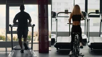 A man runs on a treadmill while a woman cycles on a stationary bike in a spacious and well-lit gym. Both are focused on their workouts, promoting an active, healthy lifestyle. video