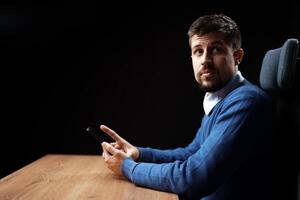 Man in blue sweater using smartphone while sitting at wooden desk in dimly lit room photo