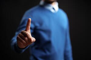 Man signaling to pause with one finger raised, dressed in casual attire against a dark background with soft lighting photo