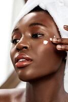 Skincare routine in a bright room featuring a woman applying moisturizer on her face with a towel on her head photo