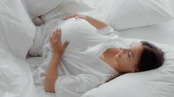 A young woman lies comfortably on her bed in a serene environment, gently cradling her pregnant belly while surrounded by soft pillows and white bedding. The atmosphere is calm and inviting. video