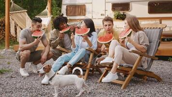 A diverse group of friends relaxes on a camping weekend in summer. They share slices of watermelon, laughter, and a dog while sitting in wooden chairs near their camper. video