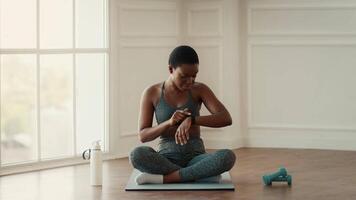A fitness enthusiast sits cross-legged on a mat, checking her fitness tracker. She is dressed in athletic wear, focused on maintaining her healthy lifestyle in a well-lit indoor space. video