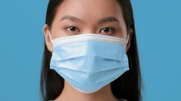 A young woman of Asian descent stands with a neutral expression against a vibrant blue background. She is wearing a medical mask, reflecting the current health climate and lifestyle choices. photo