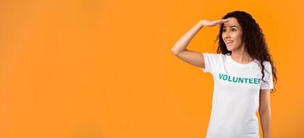 Female Volunteer Looking Aside At Free Space For Advertisement Text Holding Hand Near Eyes Posing Standing Over Yellow Studio Background. Activist Advertising Volunteering Project photo