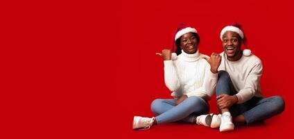 Joyful black man and woman in Santa hats pointing at copy space on orange background, demonstrating free place for your holiday advertisement or offer, cheerful african couple enjoying christmas time photo