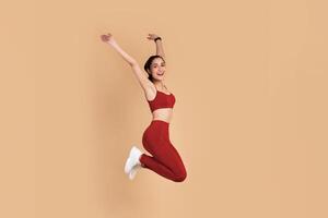 Full Length Portrait Of Happy Excited Smiling Young Woman In Sports Bra And Leggins Jumping Up And Raising Arms Up Isolated On Pastel Background, Celebrating Success, Studio Shot, Copy Space. photo
