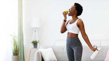 Weight Loss. Cheerful Black Lady Weighing Standing On Scales Posing Holding Apple And Water Bottle At Home. Successful Slimming And Healthy Dieting Nutrition Concept. Full Length, Empty Space photo