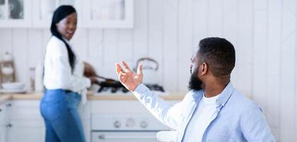 Hungry african husband complaining about empty plate, waiting for dinner in kitchen while his spouse cooking food on background, starving black man sitting at table and demanding food, free space photo