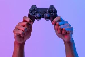 Black male hands holding joystick for games over purple background with neon lighting, unrecognizable man enjoying modern technologies for gaming, cropped image, panorama with copy space photo