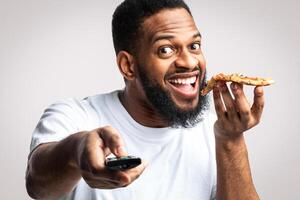 Black Man Eating Pizza And Watching TV Pointing Remote Control To Camera Posing Over White Studio Background. Junk Food Eater Watches Television Enjoying Unhealthy Fastfood. Overeating Nutrition Habit photo
