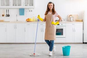Full length portrait of happy young lady with mop washing floor in kitchen, empty space. Smiling millennial maid or housewife keeping her apartment tidy and clean, doing disinfection photo