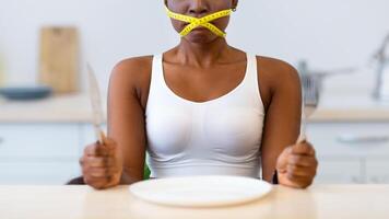 Diet Restrictions. Hungry African American Lady Having Tied Mouth With Measuring Tape Sitting Near Empty Plate, Looking At Camera In Kitchen At Home. Fasting And Weight Loss, Food Craving And Hunger photo