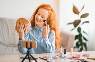 Beauty Blogging. Young beautiful red-haired teen doing makeup, recording cosmetics product review, applying powder with brush. Girl filming for her channel using mobile phone on tripod photo